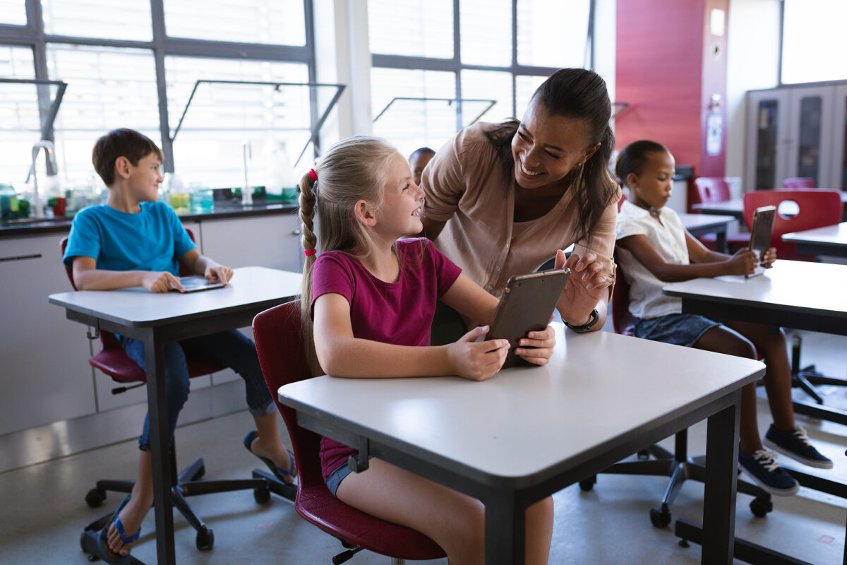 Smiling teacher helping a primary school girl use a tablet in a classroom with other children