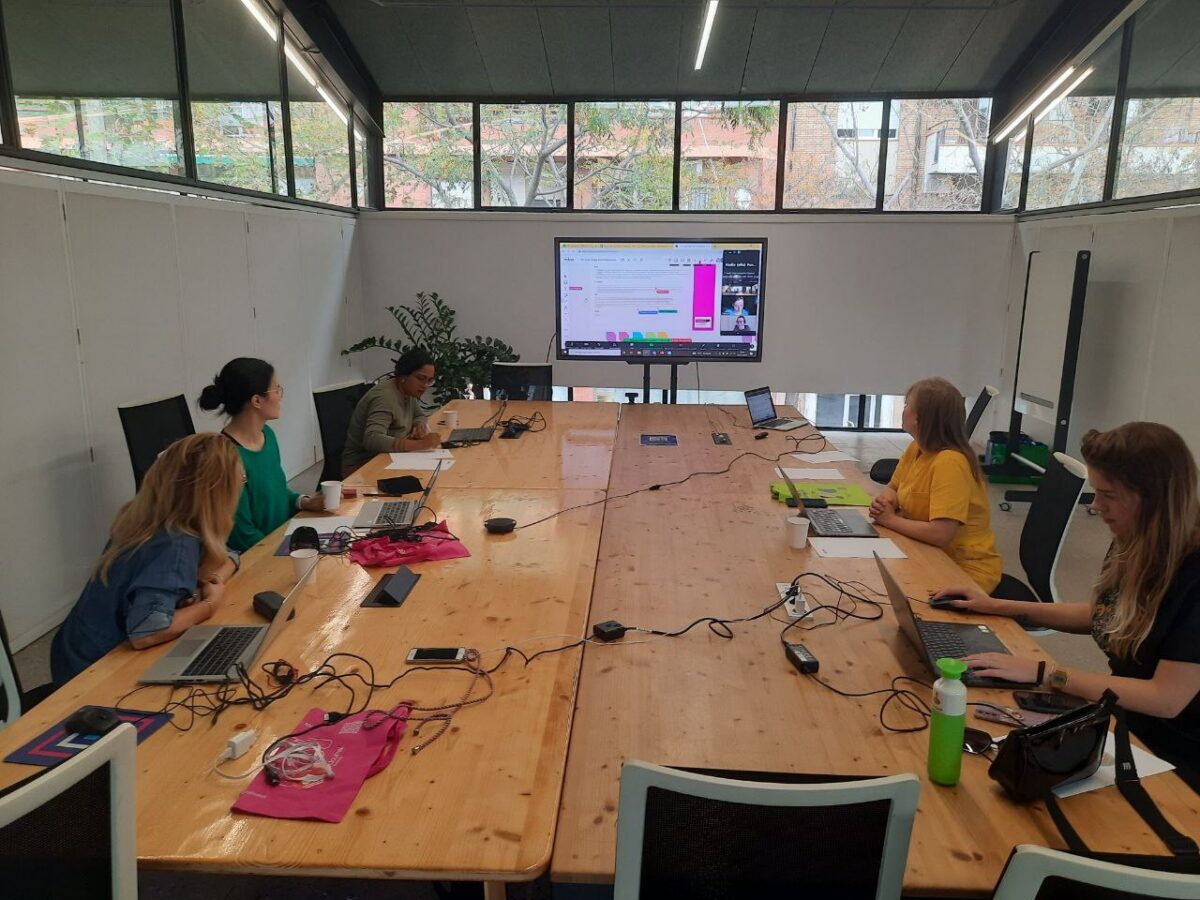 Five women sit around a large wooden table, working on laptops and participating in a video call on a screen.