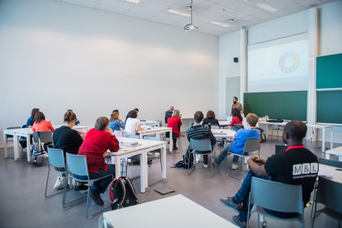 A classroom with a speaker at the front and attendees seated at tables, facing the front.