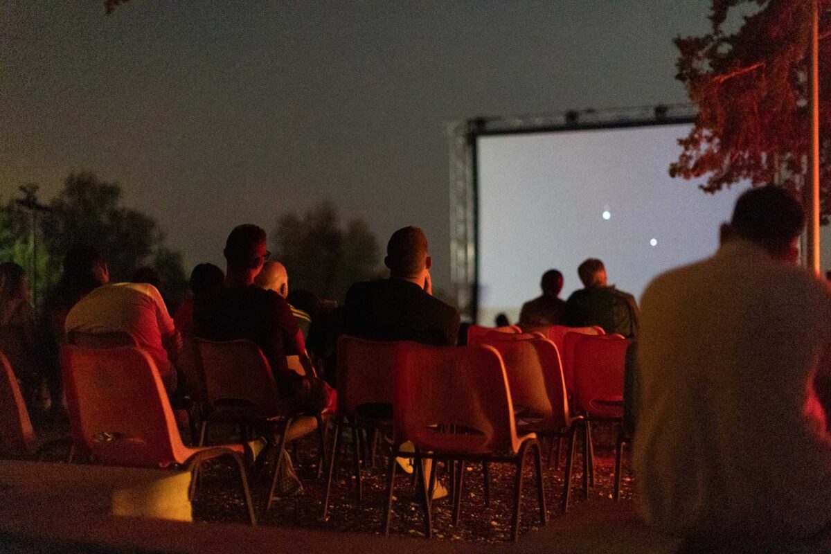 People sitting on red chairs outdoors at night, watching a film projected onto a large screen.