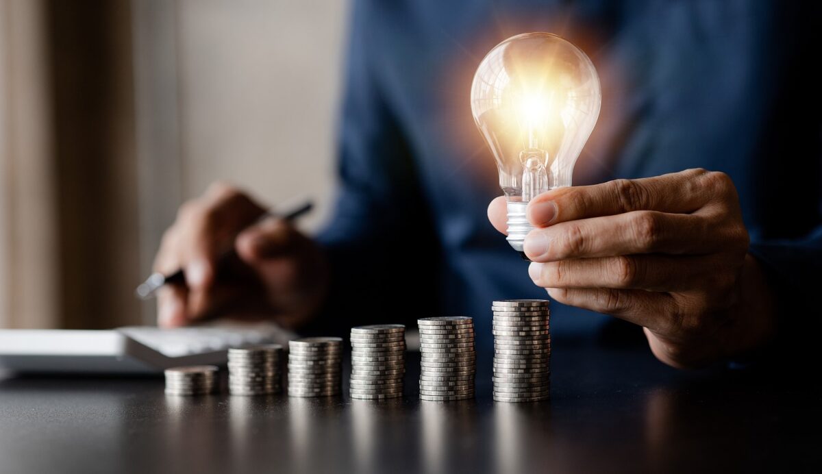 A person holds a glowing lightbulb over stacks of coins, while another person uses a calculator.