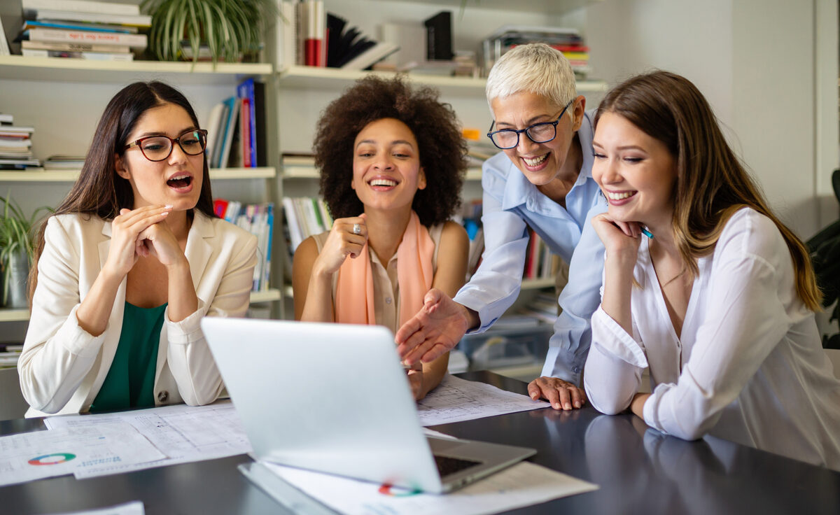 Four women, two standing and two sitting, smiling and looking at a laptop screen on a desk.