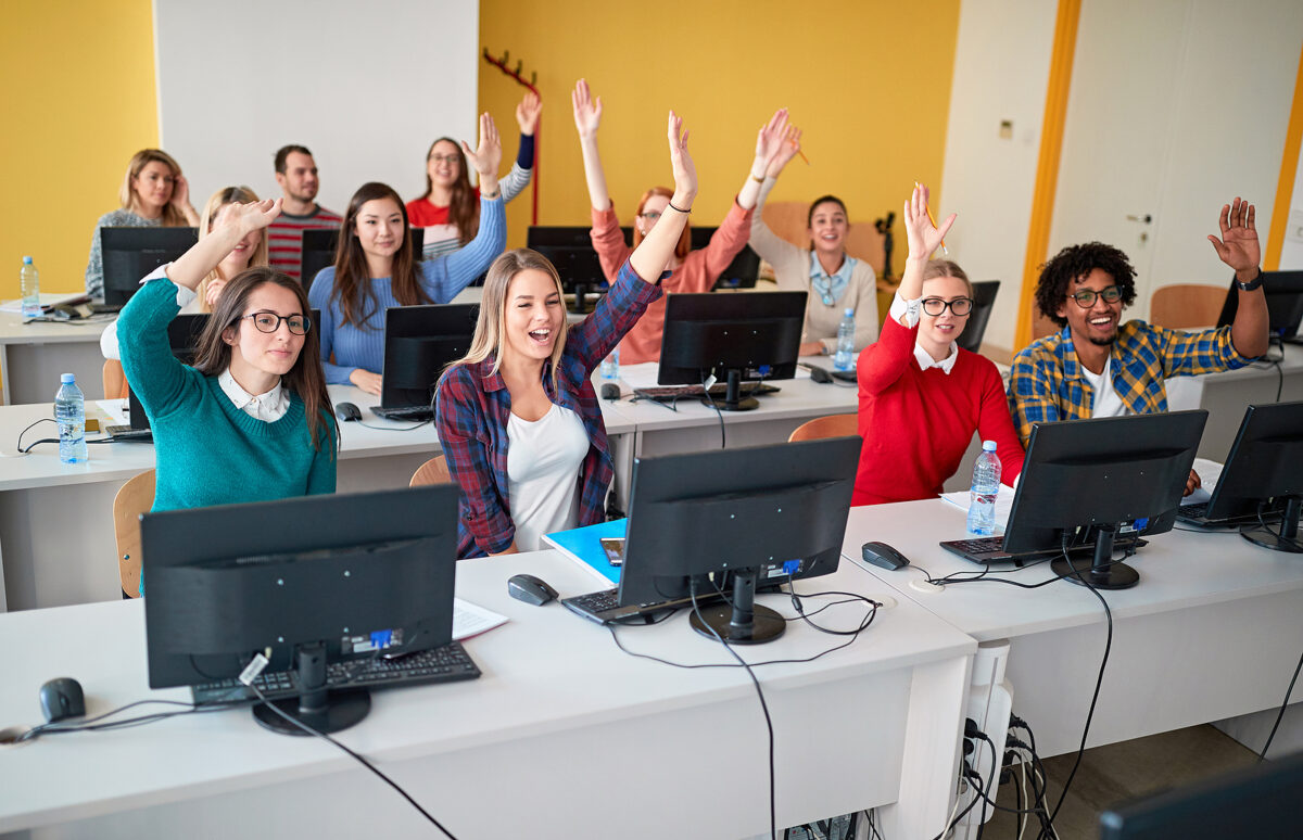 Happy students raising hands in a classroom with computers, showing engagement in a media literacy lesson.
