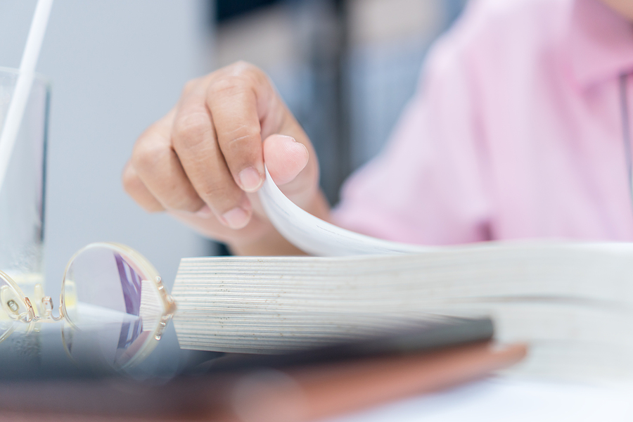 Close-up of a person's hand turning the page of a book, with spectacles and a glass nearby.