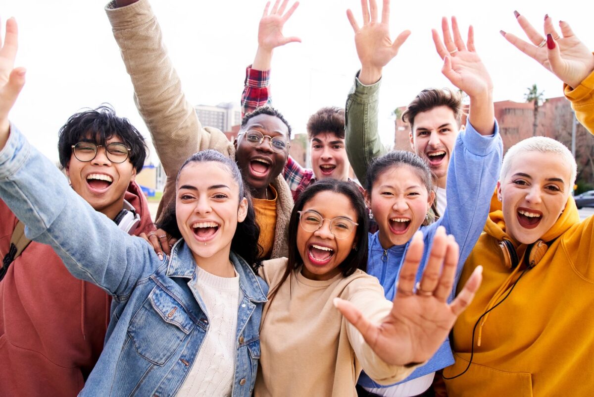 A diverse group of young people smiling and waving at the camera, with buildings in the background.
