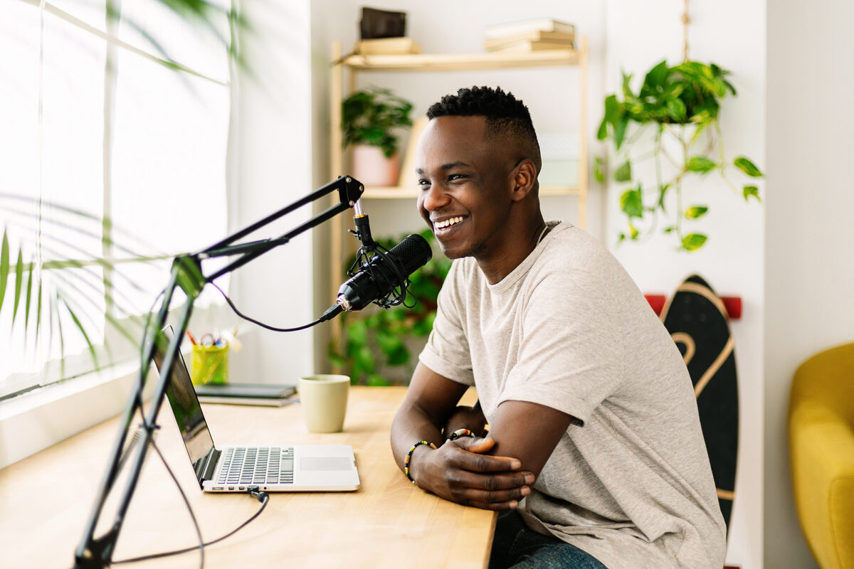 Happy young Black man recording a podcast at a desk with a laptop and microphone, smiling.