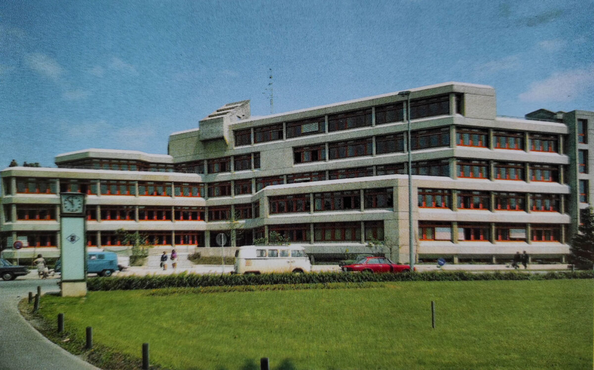A large, modern building with many windows and a clock tower in front, with a grassy area and road.