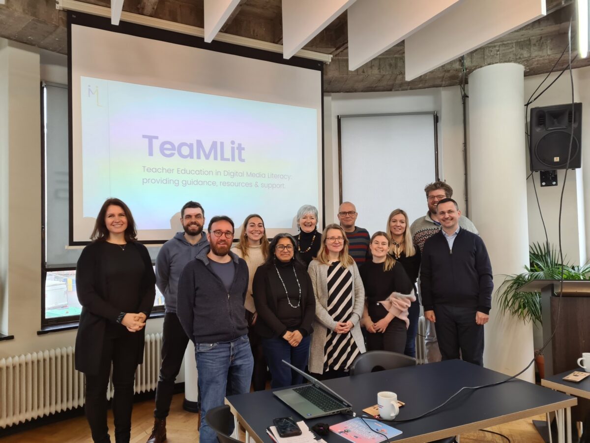 A group of 13 people, men and women, standing in front of a screen displaying the TeaMLit logo and text.
