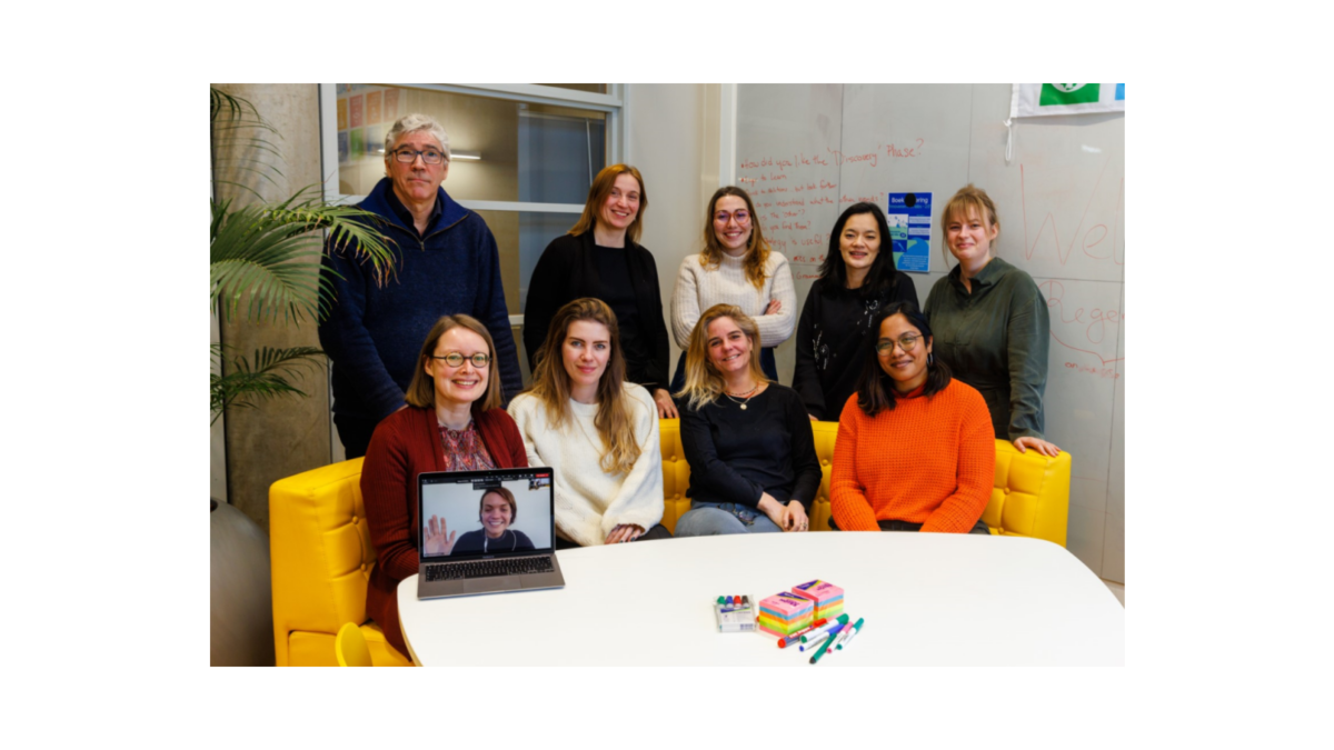 Nine people, eight women and one man, pose for a group photo in an office setting. One woman is on a laptop screen.