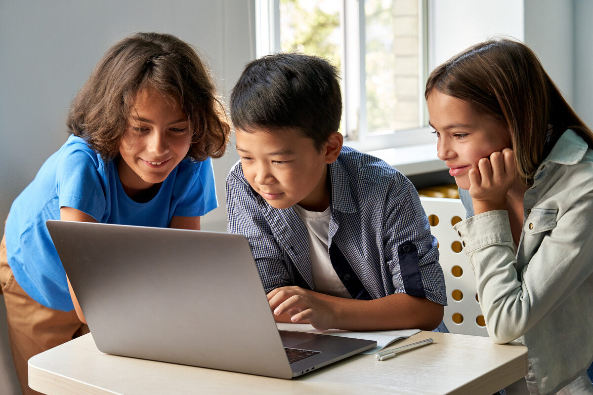 Three primary school children, two boys and one girl, looking at a laptop screen on a desk.