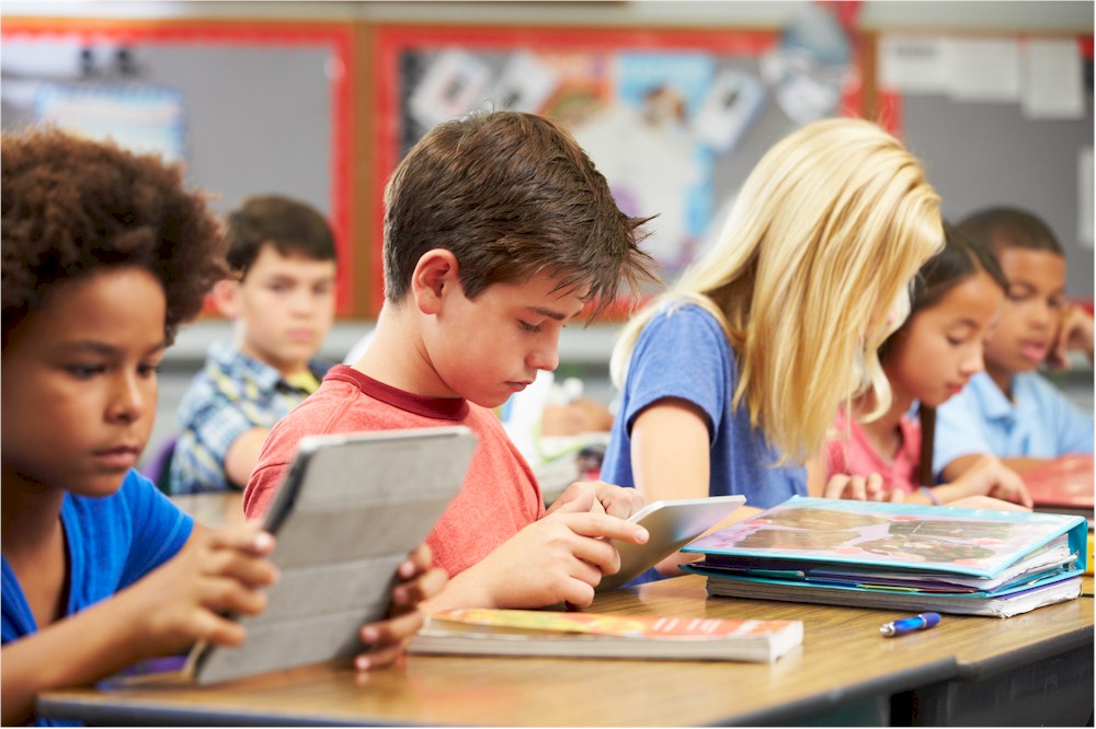 Primary school children in a classroom, some using tablets and others looking at books.