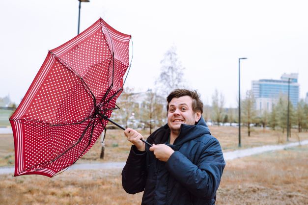 A man in a blue jacket struggles to hold an inside-out red polka dot umbrella in windy weather.