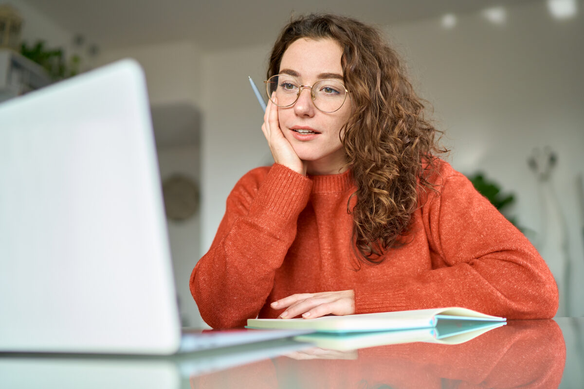 Young woman with curly hair and glasses looking at a laptop, holding a pen, with a notebook.