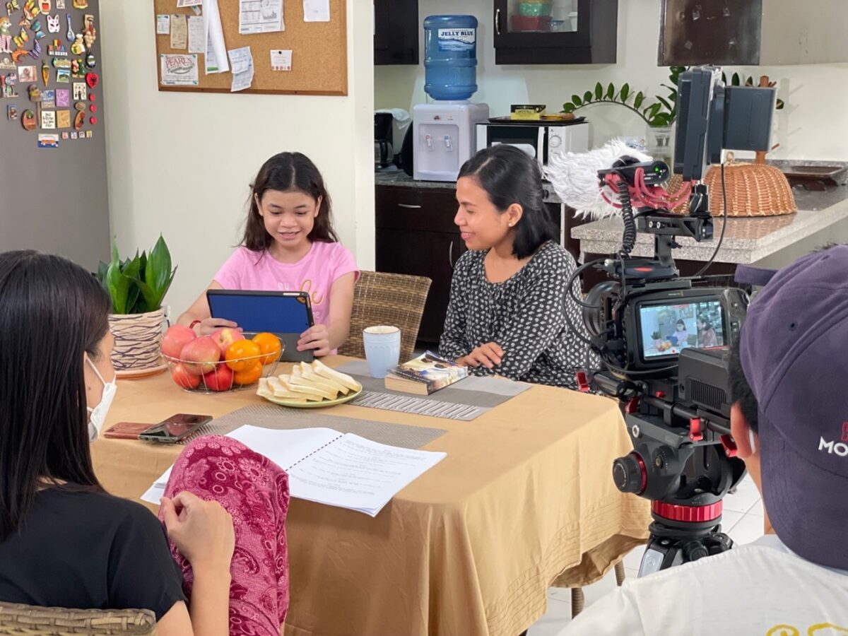 A young girl using a tablet and a woman sitting at a table, being filmed by a camera crew.