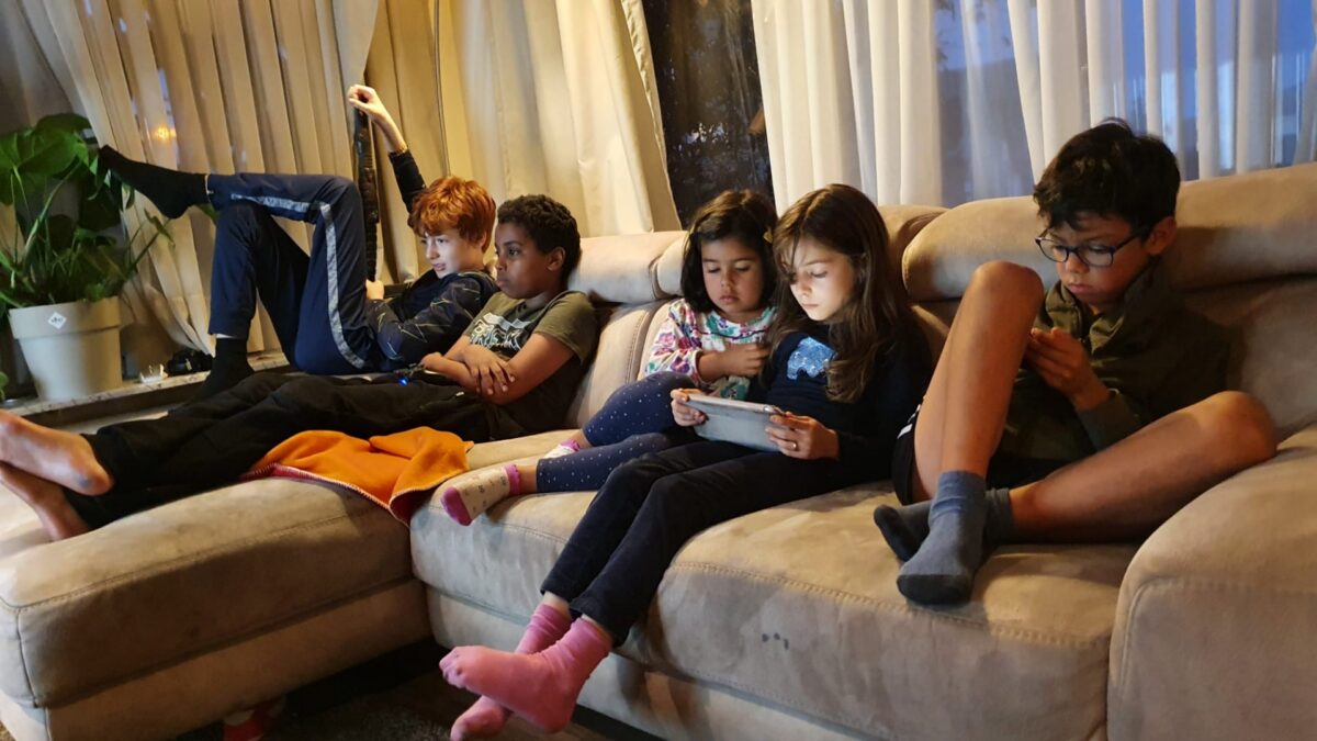 Five children and two young girls sitting on a sofa, looking at phones and tablets.