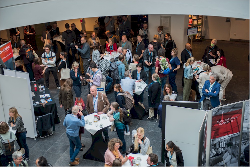 Overhead view of a crowded conference hall with attendees mingling around tables and exhibition stands.