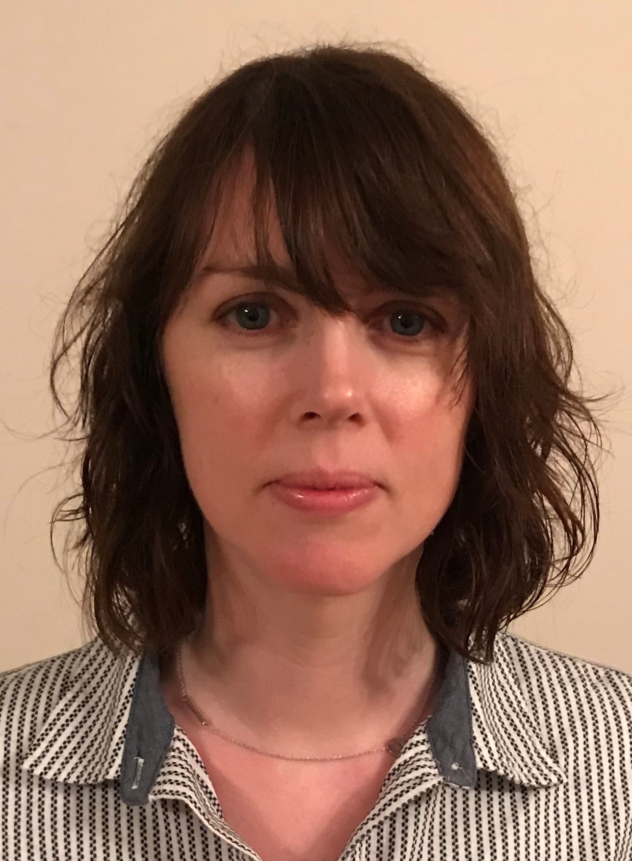 A woman with shoulder-length brown hair and bangs, wearing a striped shirt and a necklace, looking at the camera.