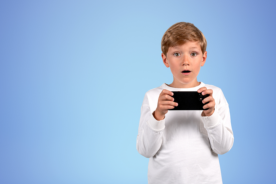 A shocked boy in a white long-sleeved shirt holds a smartphone horizontally, looking at the viewer.
