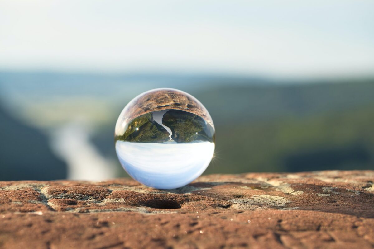 A clear glass sphere sits on a textured brown surface, reflecting an inverted landscape of green hills and a light blue sky.