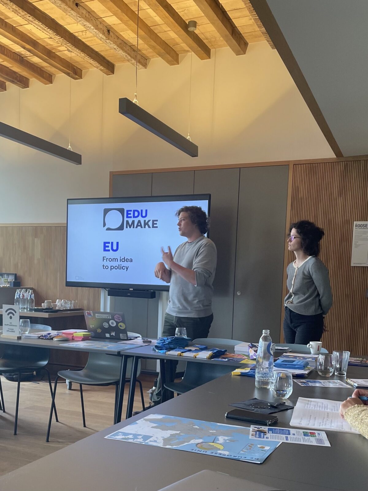 A man presenting a slide with EDU MAKE and EU From idea to policy to an audience in a conference room.