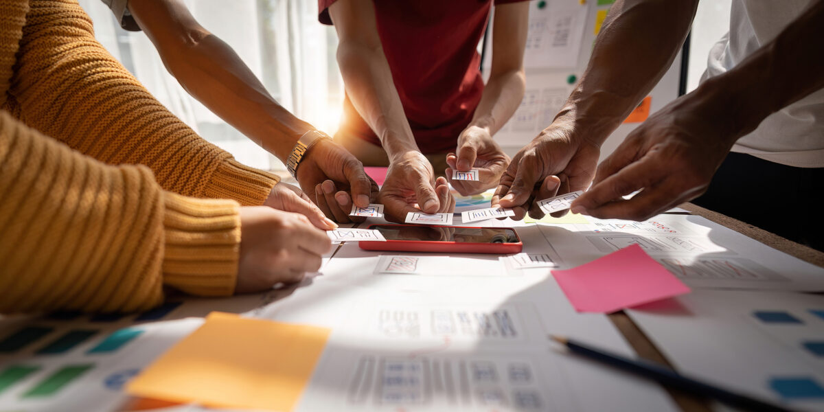 Four people's hands arranging small cards with wireframe designs around a red smartphone on a table.