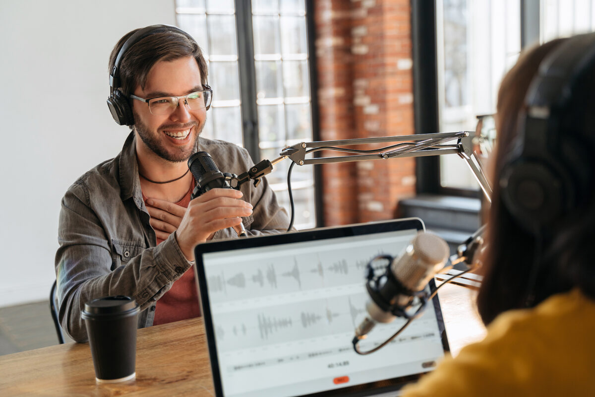 Two people with headphones and microphones recording a podcast, with a laptop displaying audio waveforms.