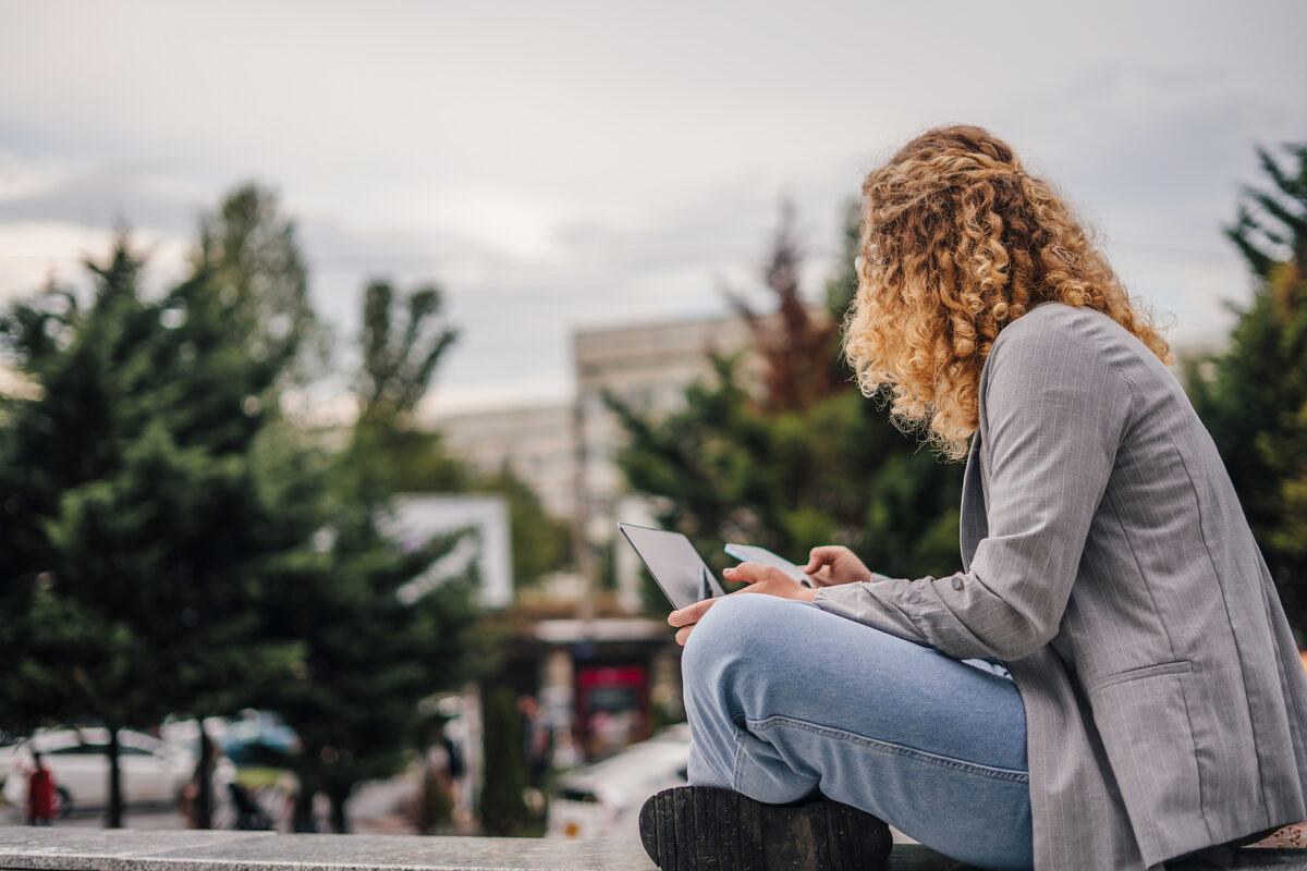 A young woman with curly blonde hair sits outdoors, using a tablet and a smartphone.