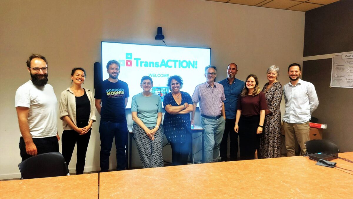 Eleven people, five women and six men, stand in a line in front of a screen displaying the text TransACTION!