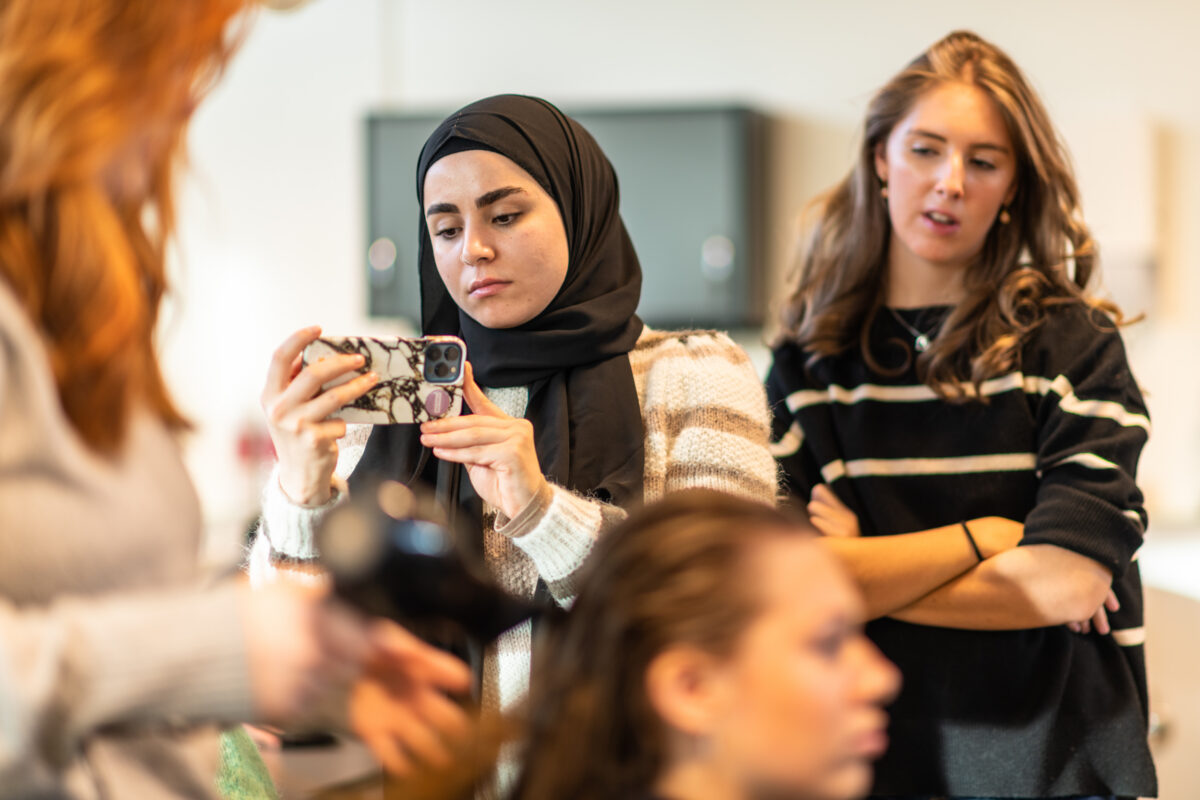 A young woman in a hijab films another woman having her hair styled, while a third woman watches.