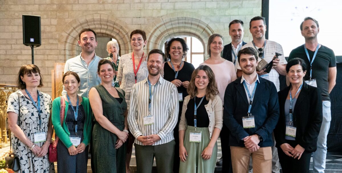 Group photo of 15 people, some smiling, in a hall with stone arches and a speaker on a stand.