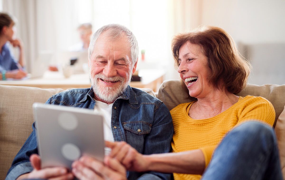 An older man and woman sitting on a sofa, looking at a tablet and laughing. The man is pointing at the screen.
