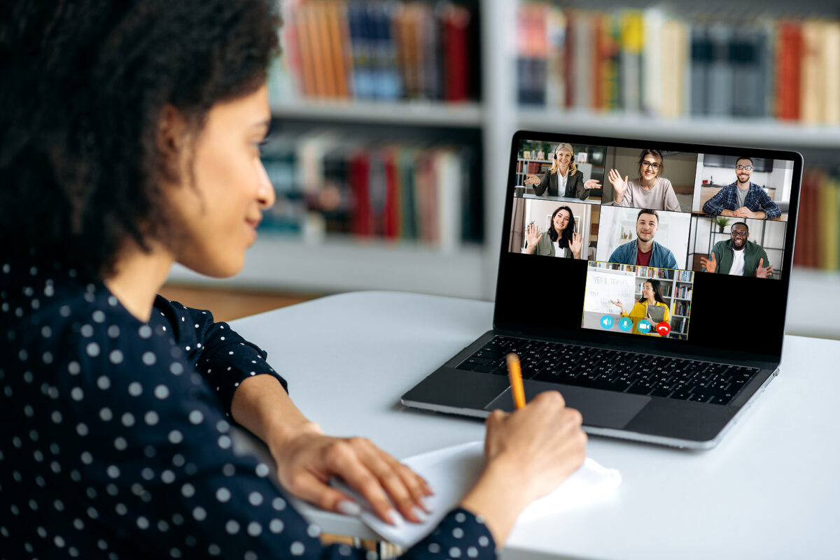 A woman takes notes while participating in a video conference with six other people on her laptop.