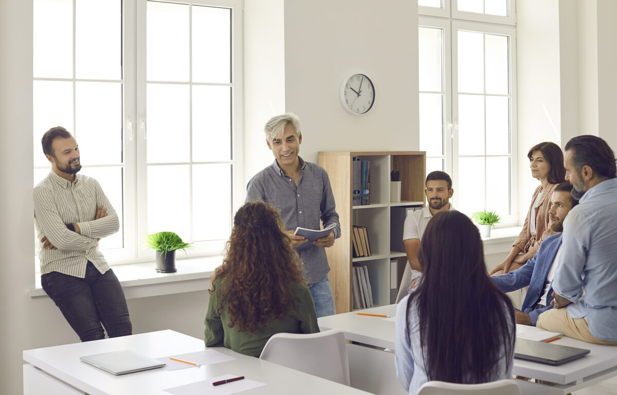 A group of adults in a bright room, some sitting at desks, others standing, listening to a man.