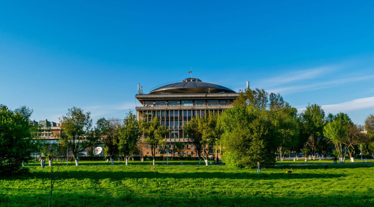 A large, modern building with a circular, domed roof, surrounded by green trees and grass.