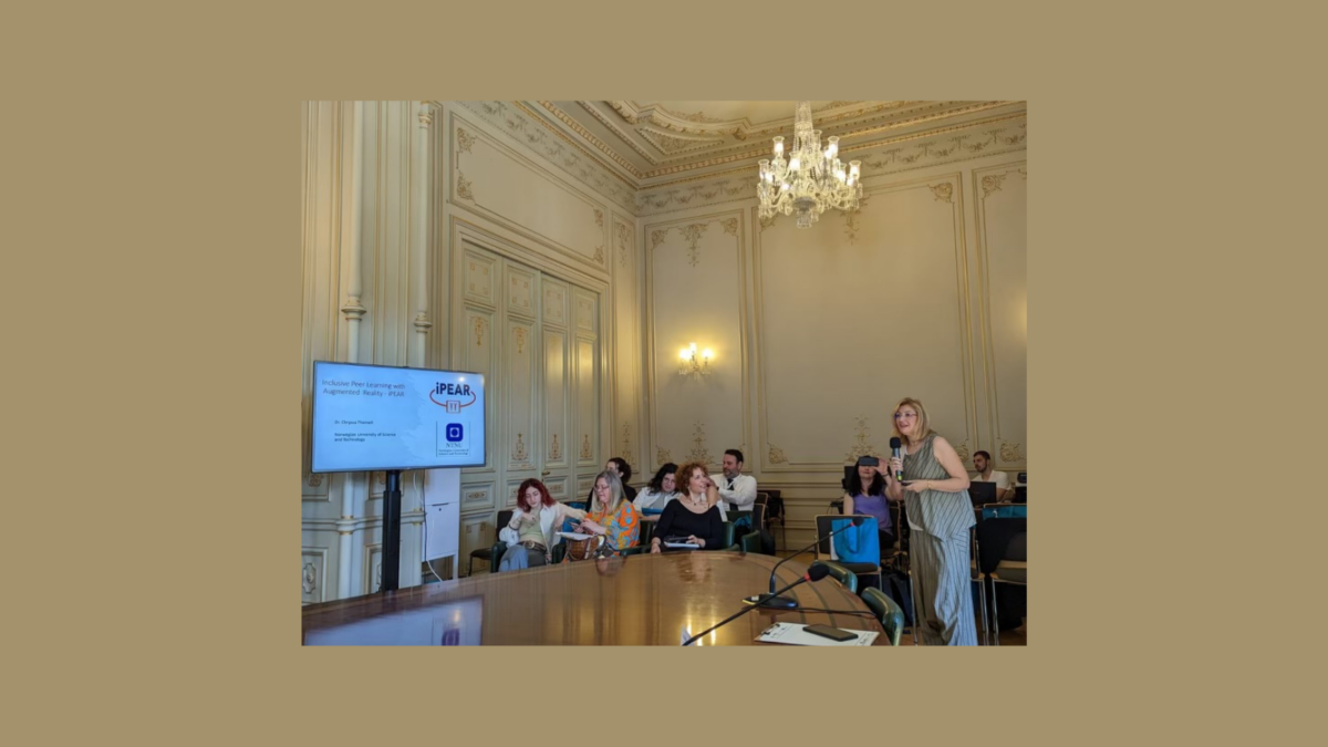 A woman speaking into a microphone at a conference, with attendees seated at a large table and a screen displaying a