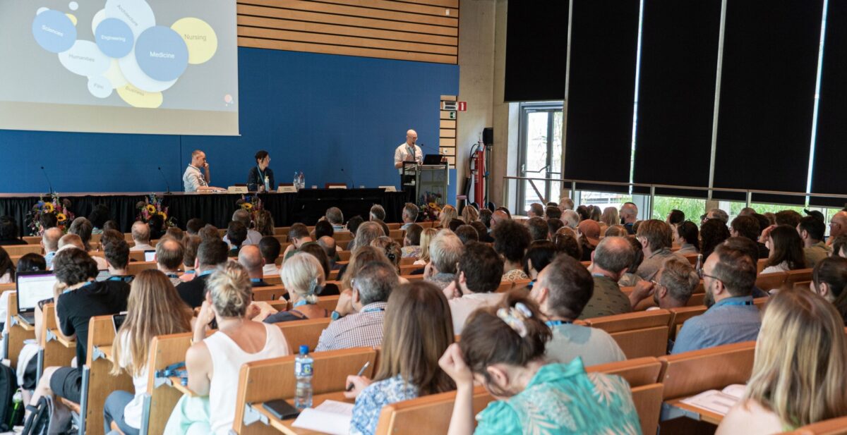 A speaker at a podium presenting to a large audience in a lecture hall, with a screen showing a diagram.