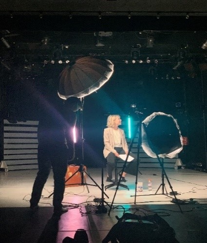 A woman sits on a stool on a stage, holding papers, with a person and lighting equipment around her.