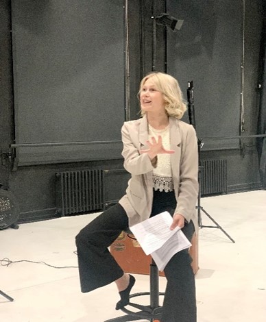 A woman with blonde hair, wearing a beige blazer and black trousers, sits on a stool, gesturing with one hand and holding