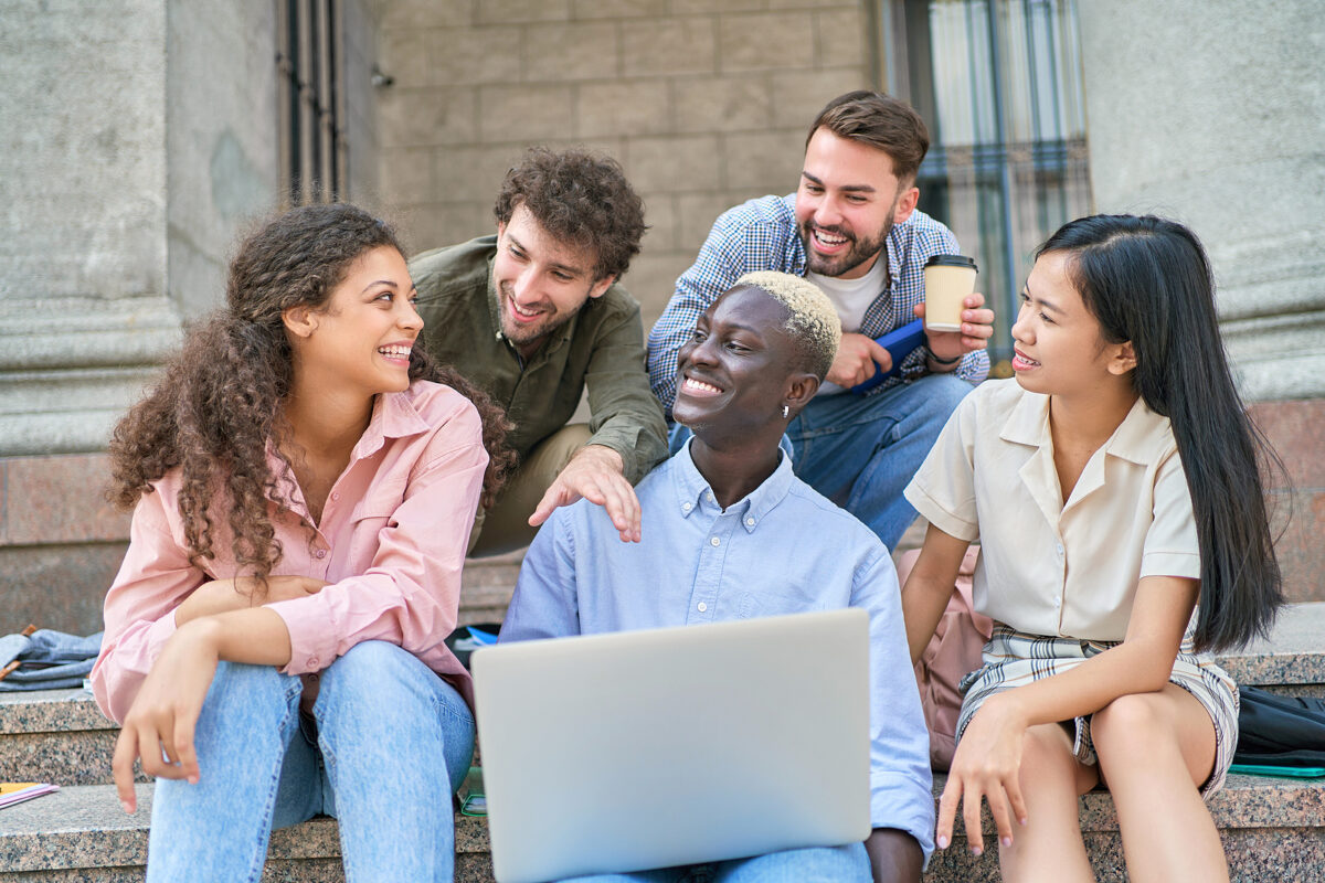 Five smiling university students of diverse backgrounds sitting on steps, with one using a laptop.