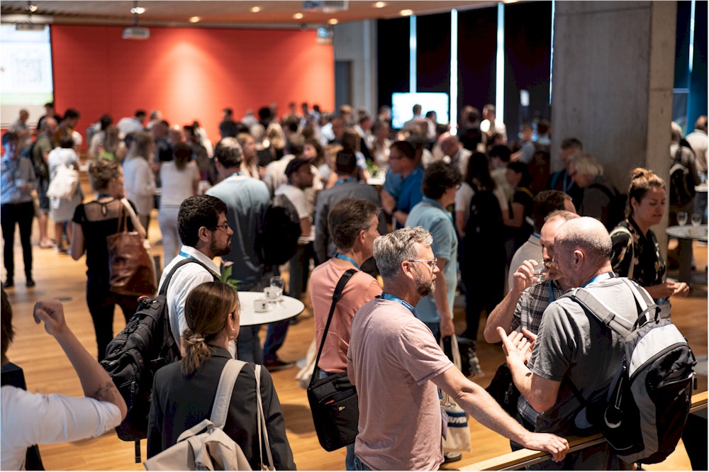 A large group of people mingling and talking in a conference hall with a red wall and screens.