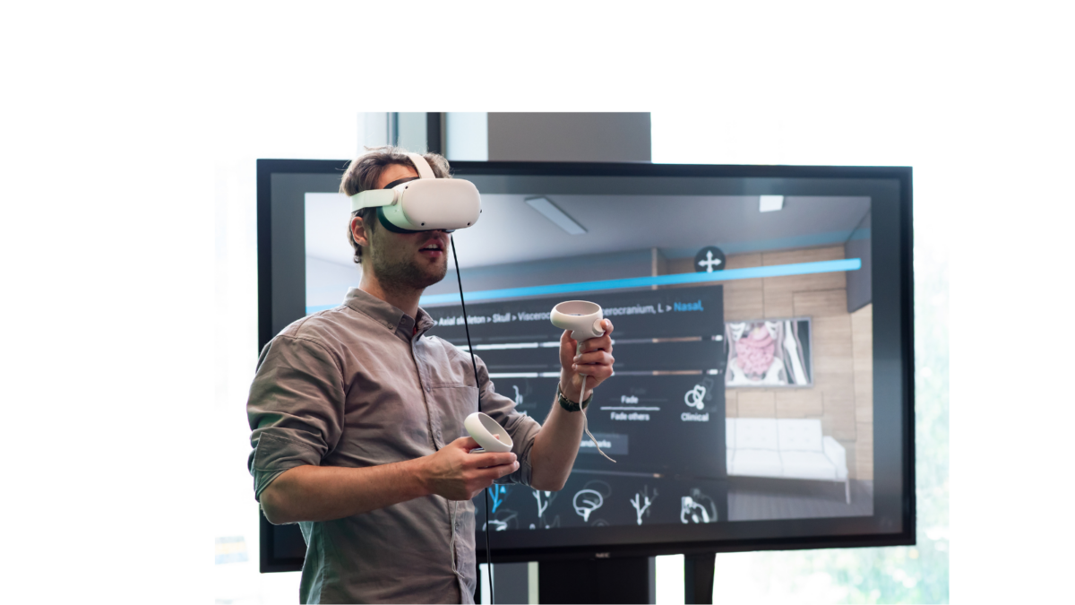 A man wearing a white VR headset and holding two white controllers, standing in front of a large screen