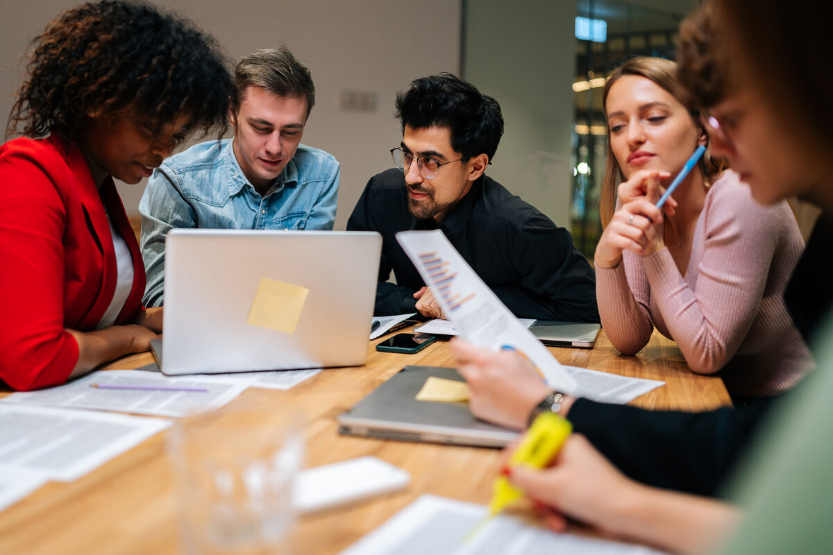 Five people around a table looking at a laptop and documents, one person highlighting text.