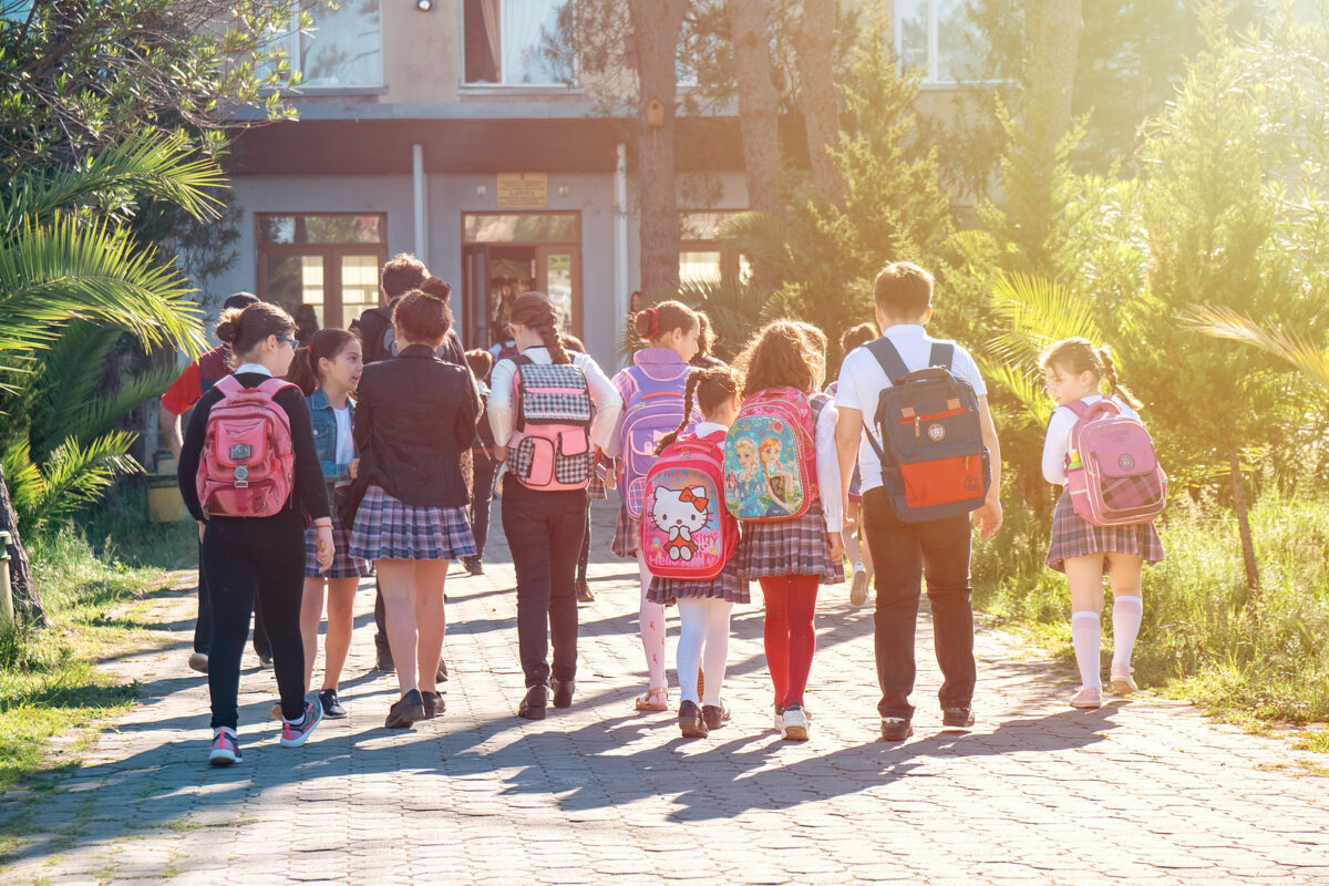 A group of school children with backpacks walking away from the camera towards a school building.