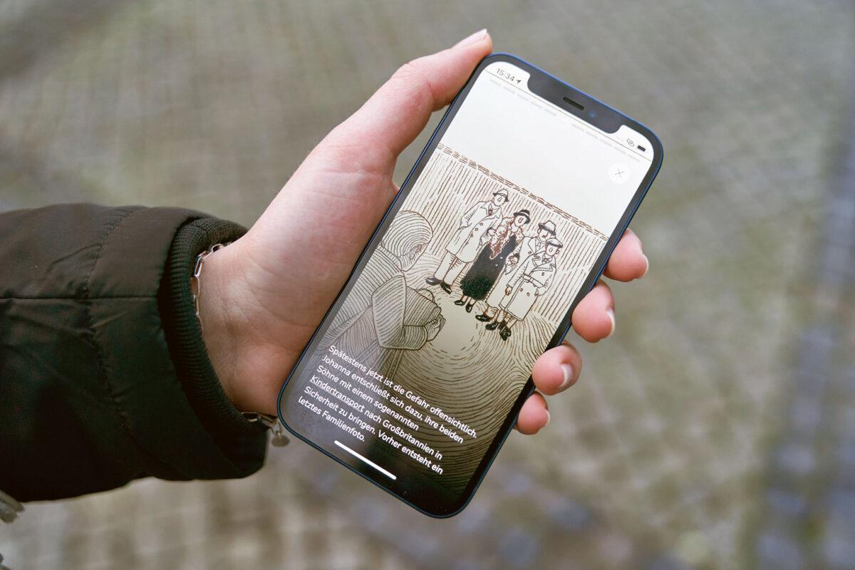 A person holds a smartphone displaying a black and white illustration of people and German text.