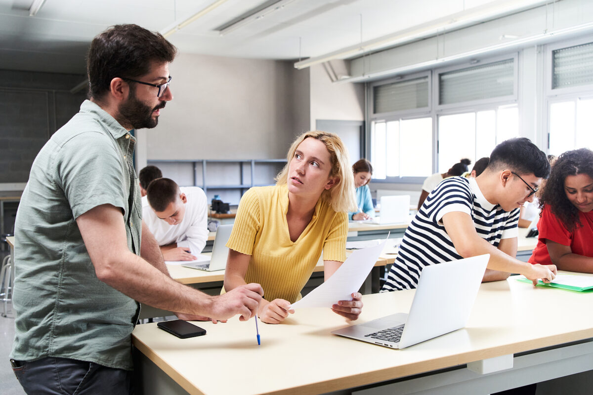 A male teacher with a beard and glasses helps a female student with a laptop and papers in a classroom.