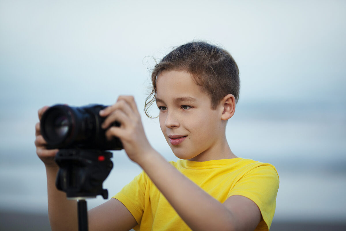 A boy in a yellow t-shirt adjusts a digital camera mounted on a tripod, looking at the screen.