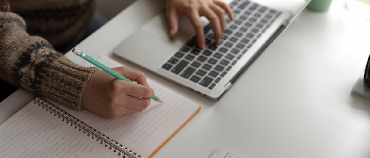 A student in a patterned jumper takes notes with a pencil in a spiral notebook while typing on a laptop.