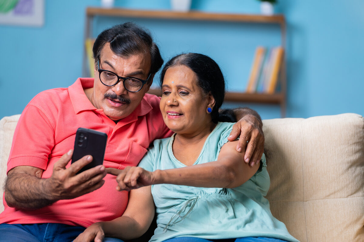 Happy Indian senior couple sitting on a sofa, looking at and pointing to a smartphone screen.