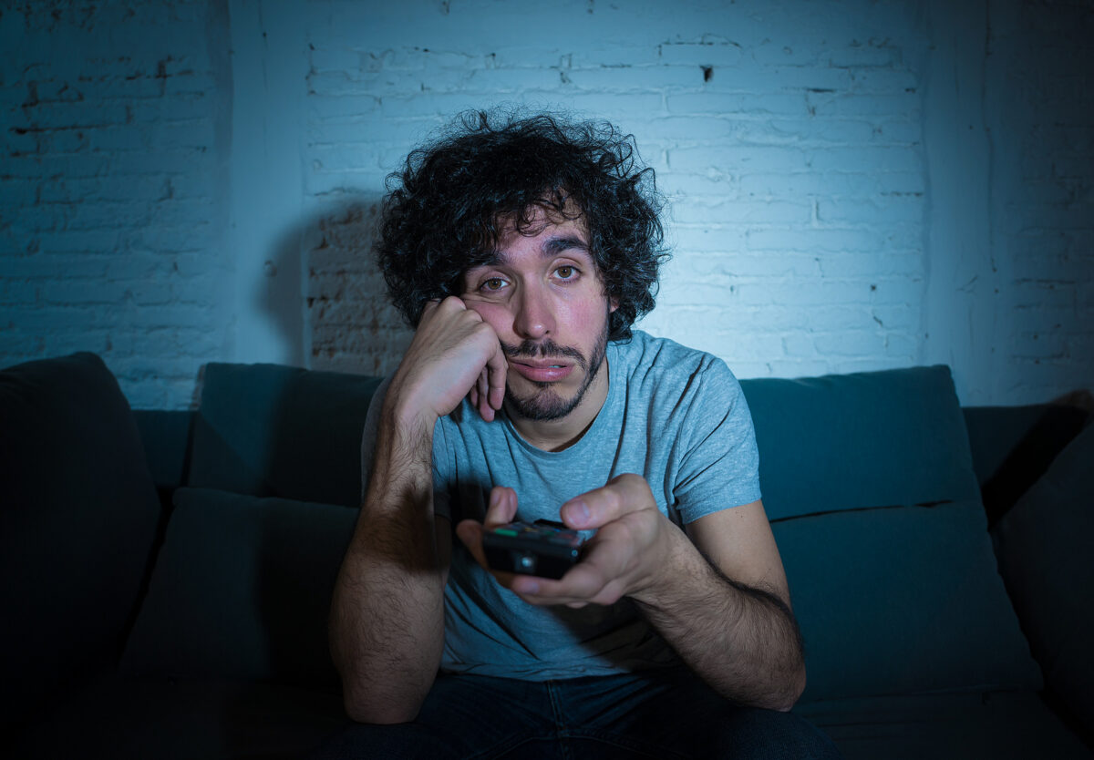 A young man with curly hair, looking bored and tired, holding a TV remote control.