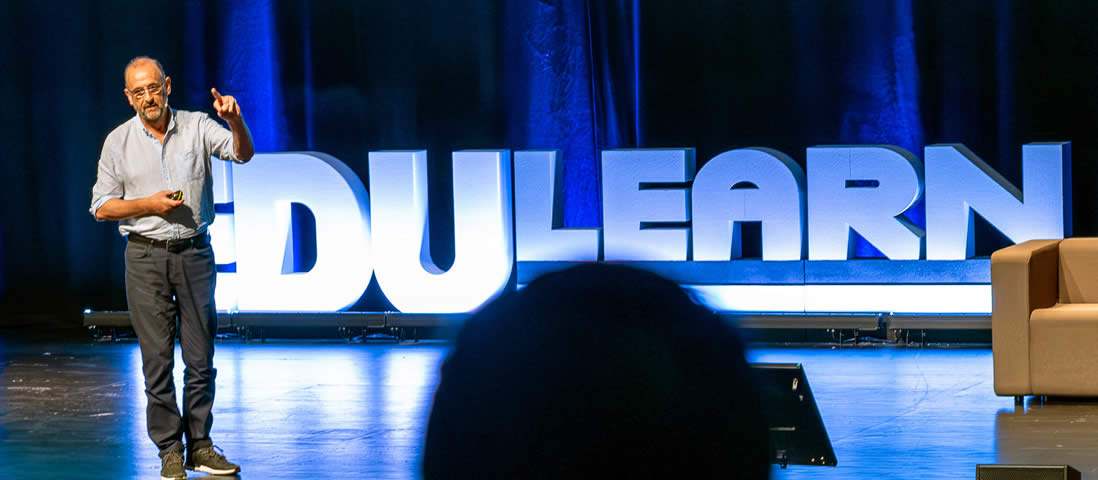Speaker on stage at EDULEARN conference, pointing, with large white EDULEARN letters in background